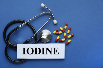 Photo of Card with word Iodine, stethoscope and pills on blue background, flat lay