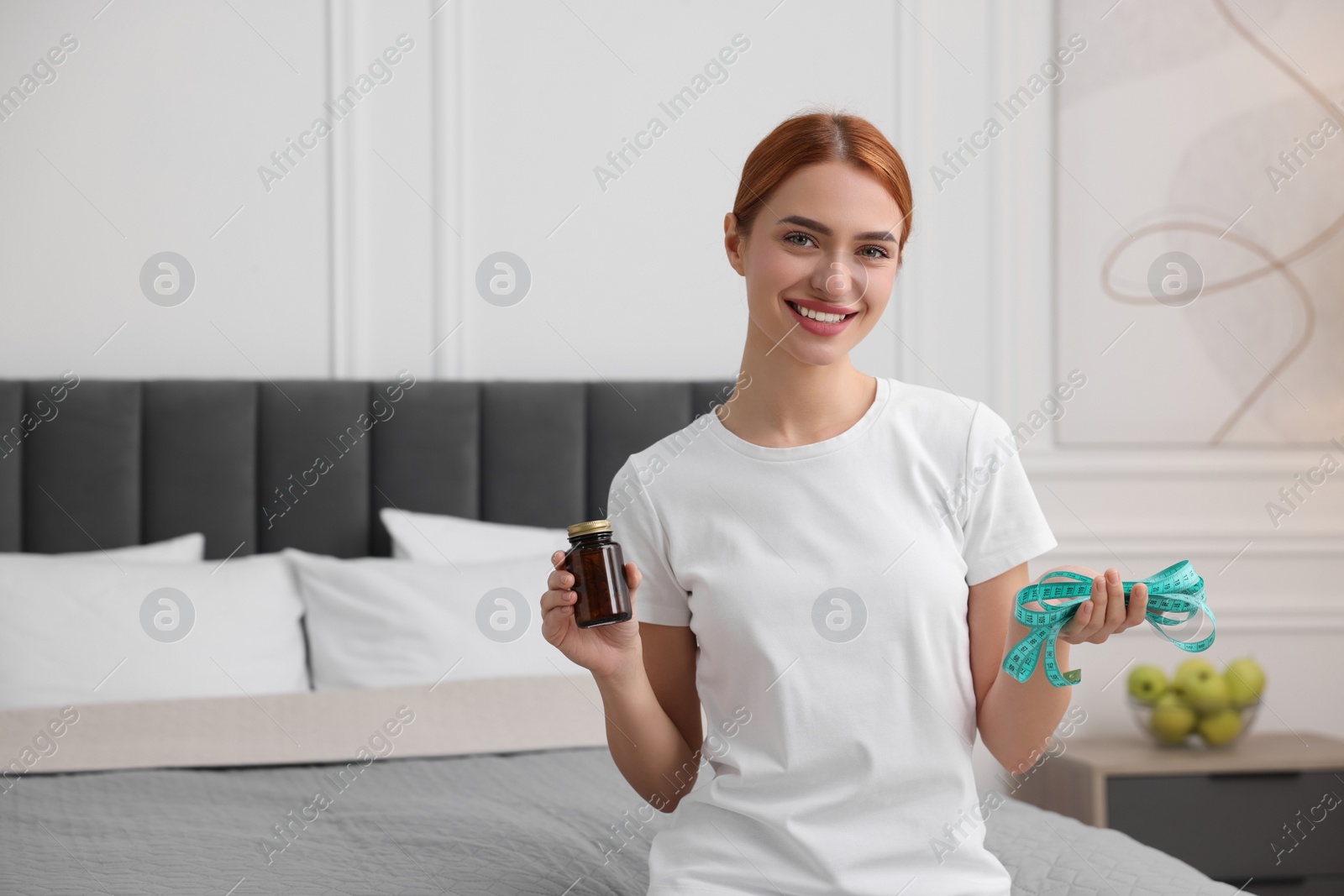 Photo of Happy young woman with pills and measuring tape on bed in room, space for text. Weight loss