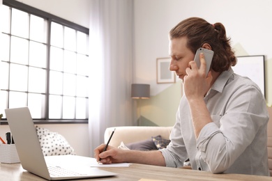 Young man talking on phone while working with laptop at desk in home office