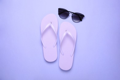 Photo of Stylish flip flops and sunglasses on light purple background, flat lay
