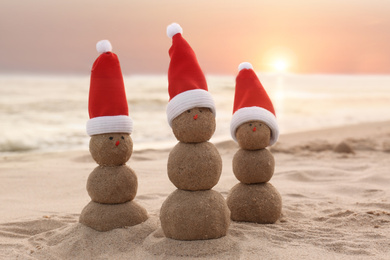 Photo of Snowmen made of sand with Santa hats on beach near sea at sunset. Christmas vacation