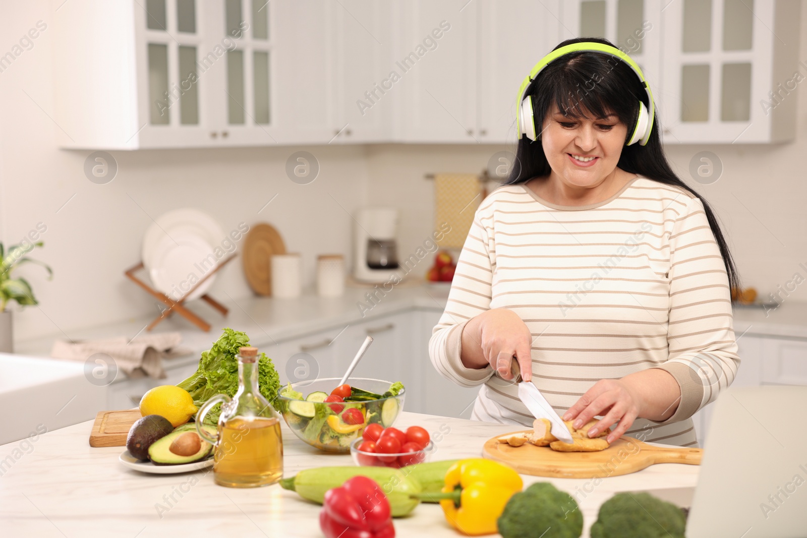 Photo of Happy overweight woman with headphones preparing healthy meal at table in kitchen