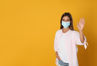 Photo of Woman in protective mask showing hello gesture on yellow background, space for text. Keeping social distance during coronavirus pandemic