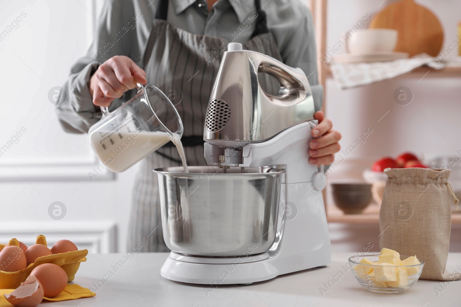 Photo of Woman pouring milk into bowl of stand mixer while making dough at table indoors, closeup