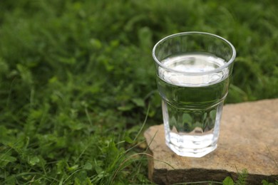 Photo of Glass of fresh water on stone in green grass outdoors. Space for text