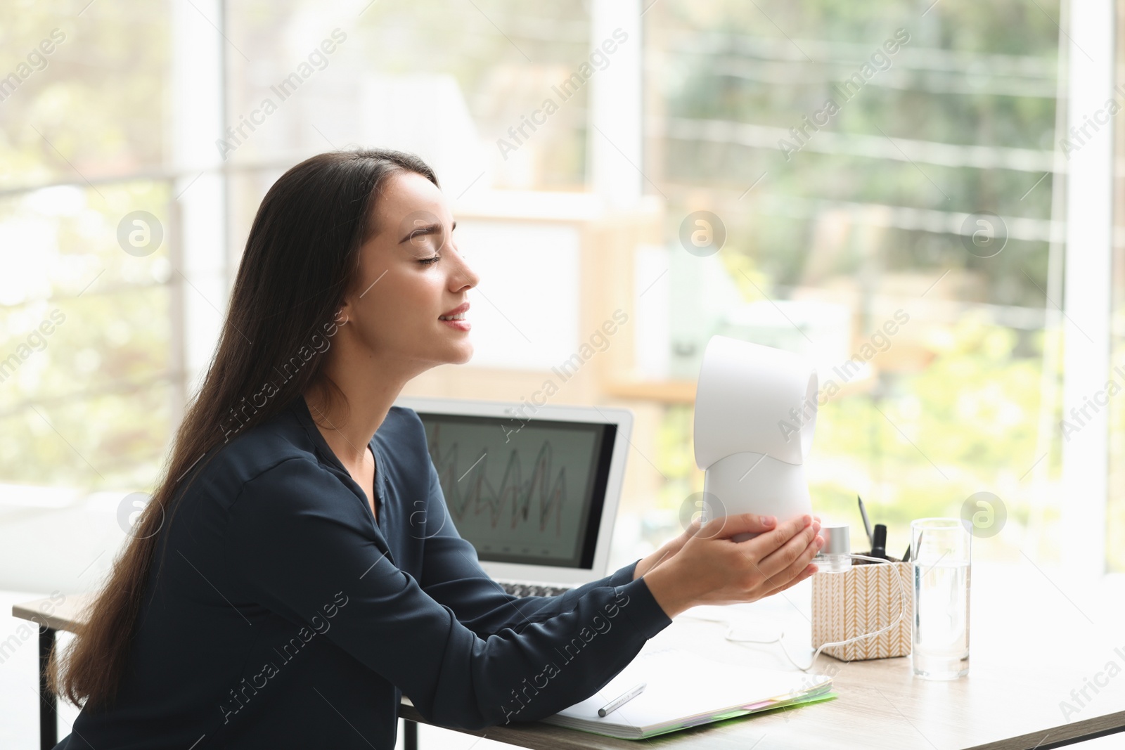 Photo of Young woman enjoying air flow from portable fan at workplace. Summer heat