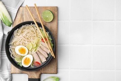Photo of Bowl of delicious rice noodle soup with meat and egg on white tiled table, flat lay. Space for text