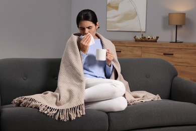 Photo of Woman with tissue and cup of drink coughing on sofa at home. Cold symptoms