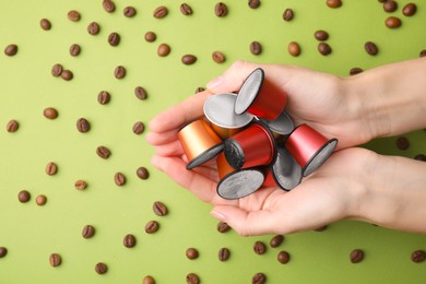 Woman holding heap of coffee capsules over beans on green background, top view