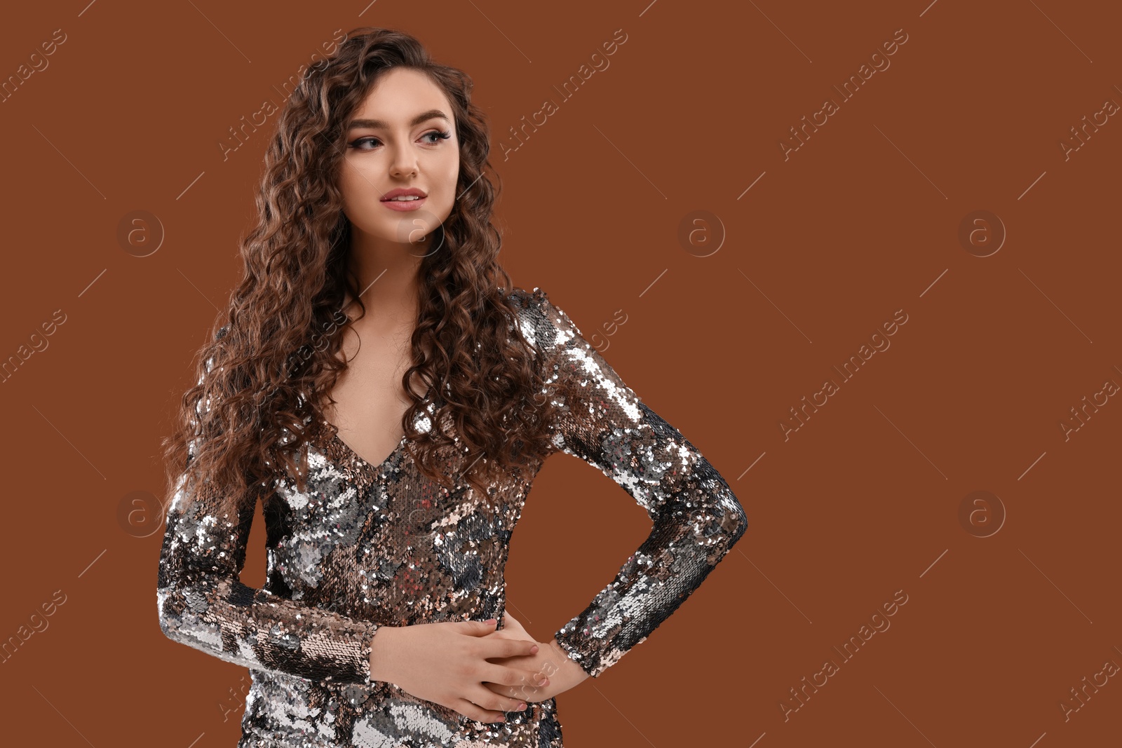 Photo of Beautiful young woman with long curly hair in sequin dress on brown background, space for text