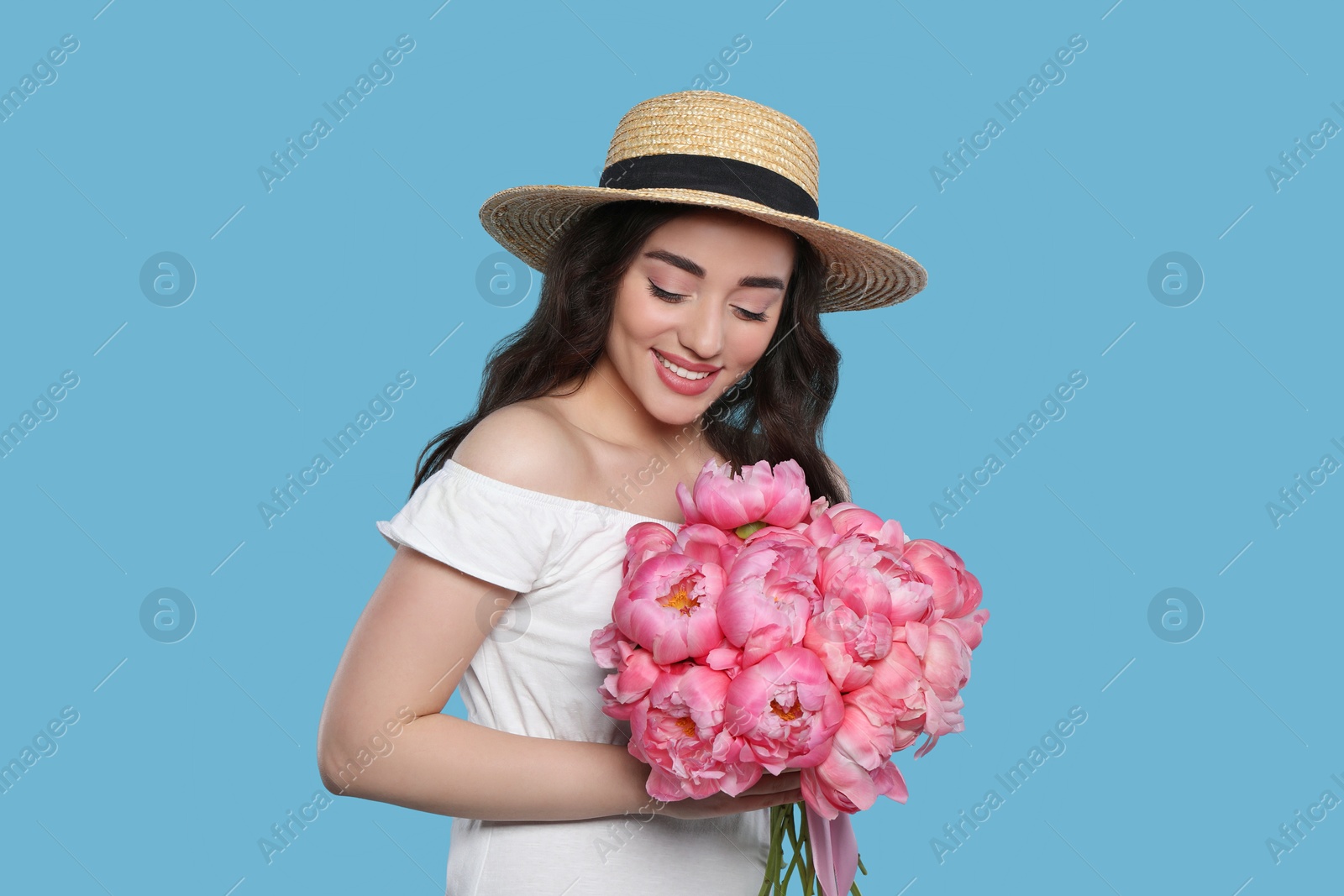 Photo of Beautiful young woman in straw hat with bouquet of pink peonies against light blue background