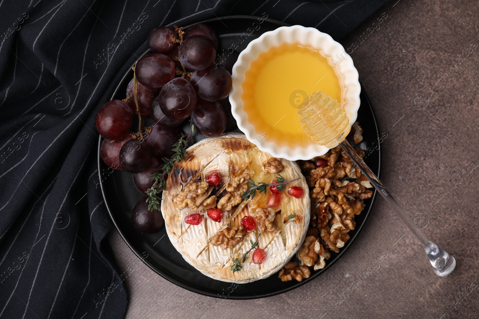 Photo of Plate with tasty baked camembert, honey, grapes, walnuts and pomegranate seeds on brown textured table, top view