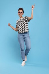 Happy young woman in sunglasses dancing on light blue background