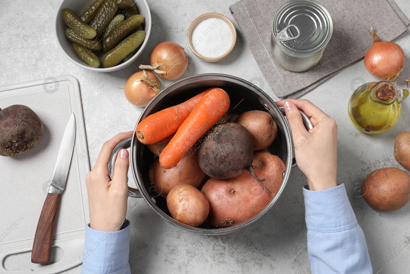 Photo of Woman holding pot with fresh vegetables at white table, top view. Cooking vinaigrette salad