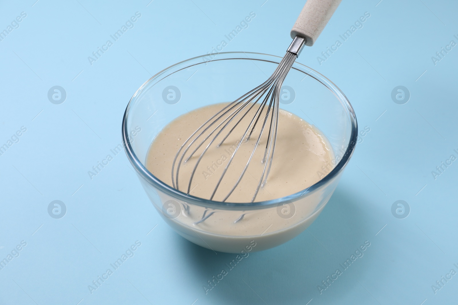 Photo of Dough and whisk in glass bowl on light blue background