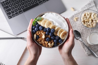 Woman holding bowl of tasty granola over white wooden table with laptop, top view