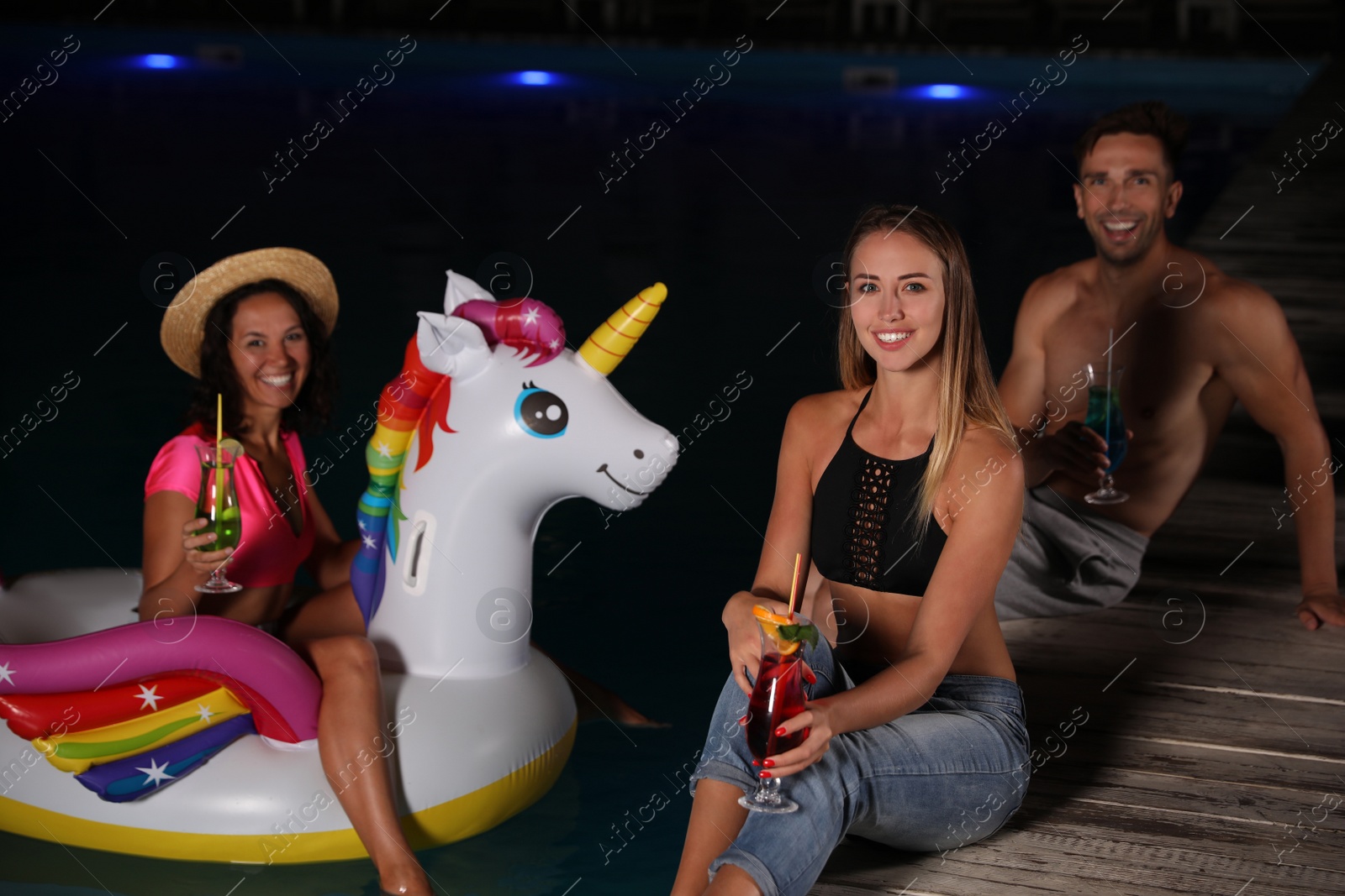 Photo of Happy young friends with refreshing cocktails relaxing near outdoor swimming pool at night