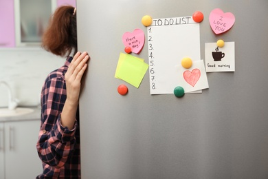 Photo of Woman opening refrigerator door with paper sheets and magnets at home, closeup