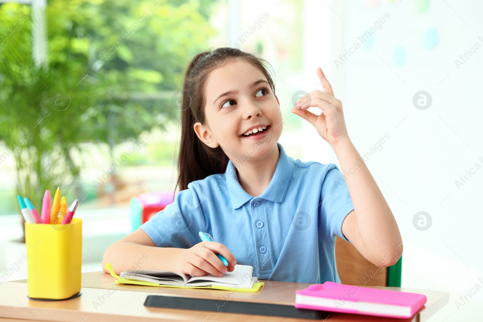 Photo of Smart little girl doing assignment at desk in classroom. School stationery