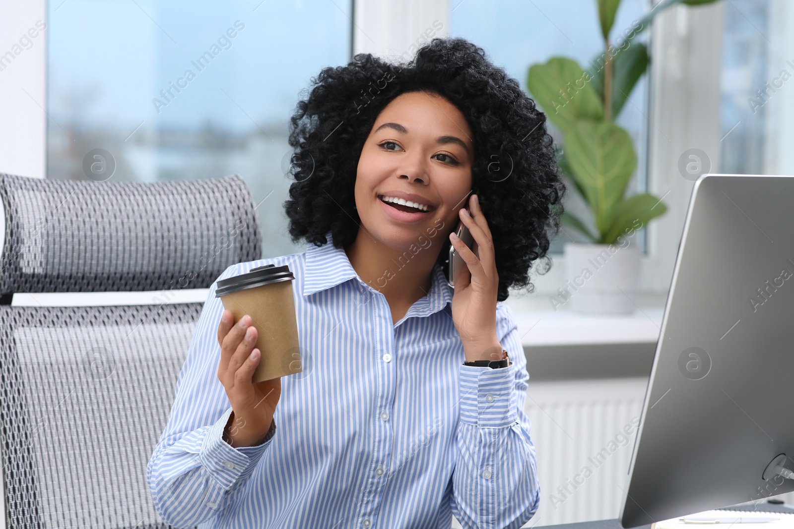 Photo of Young woman with cup of drink talking on phone in office