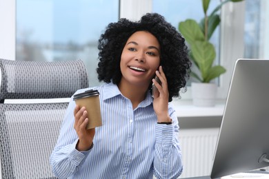 Photo of Young woman with cup of drink talking on phone in office