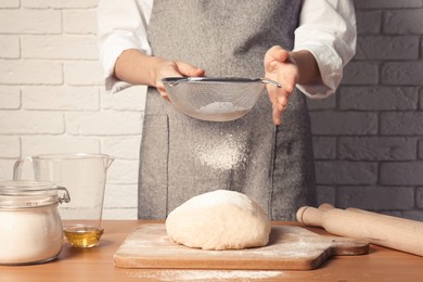 Woman sprinkling flour over dough at table near white brick wall, closeup