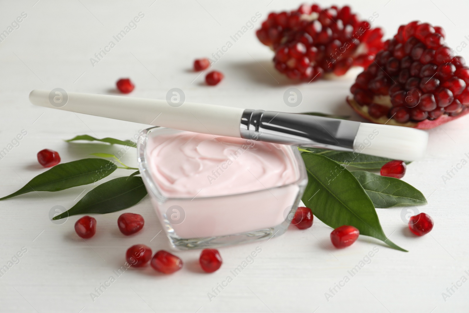 Photo of Glass bowl with natural facial mask, pomegranate seeds and brush on white wooden table, closeup