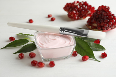 Photo of Glass bowl with natural facial mask, pomegranate seeds and brush on white wooden table, closeup