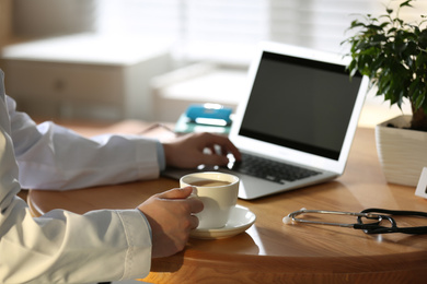 Professional doctor with cup of coffee working on laptop in office, closeup