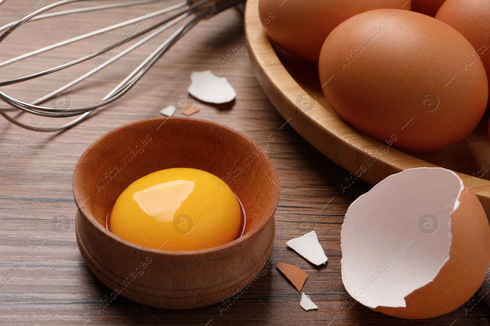 Photo of Chicken eggs, bowl with raw yolk and whisk on wooden table, closeup