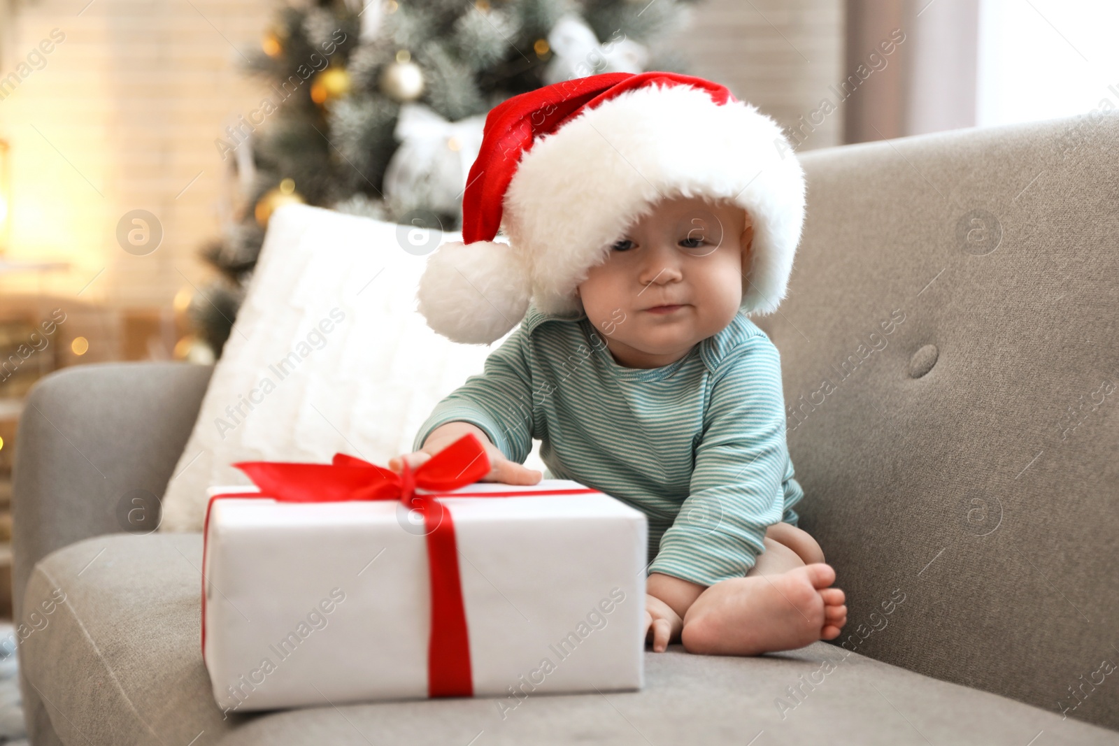 Image of Cute baby in Santa hat with Christmas gift sitting on sofa at home