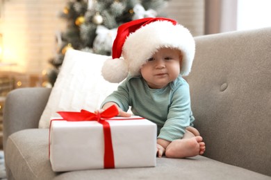 Image of Cute baby in Santa hat with Christmas gift sitting on sofa at home