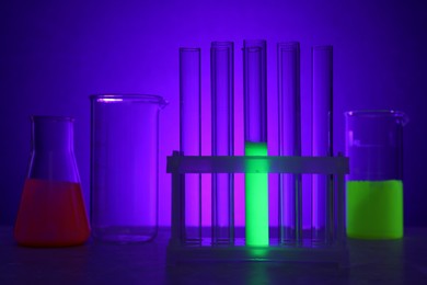 Laboratory glassware with luminous liquids on table against purple background
