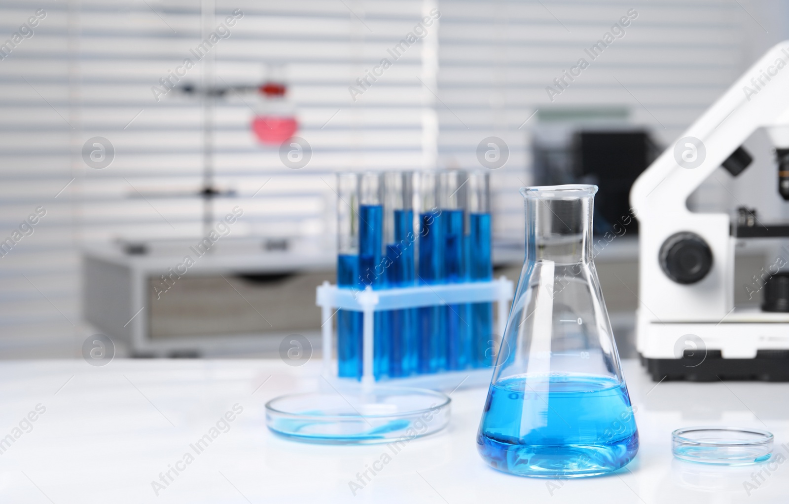 Photo of Laboratory analysis. Different glassware with blue liquid and microscope on white table indoors, space for text