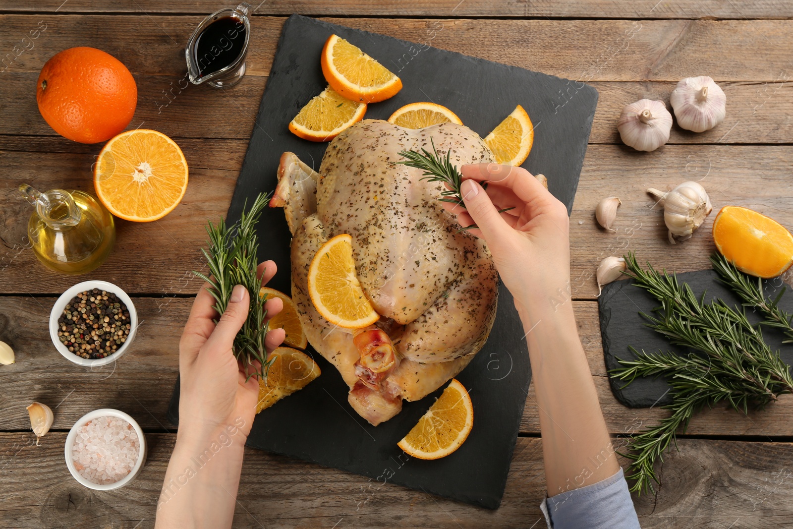 Photo of Woman adding rosemary to raw chicken with orange slices at wooden table, top view