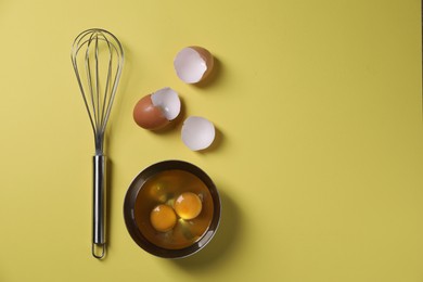 Photo of Metal whisk, raw eggs in bowl and shells on yellow background, flat lay. Space for text