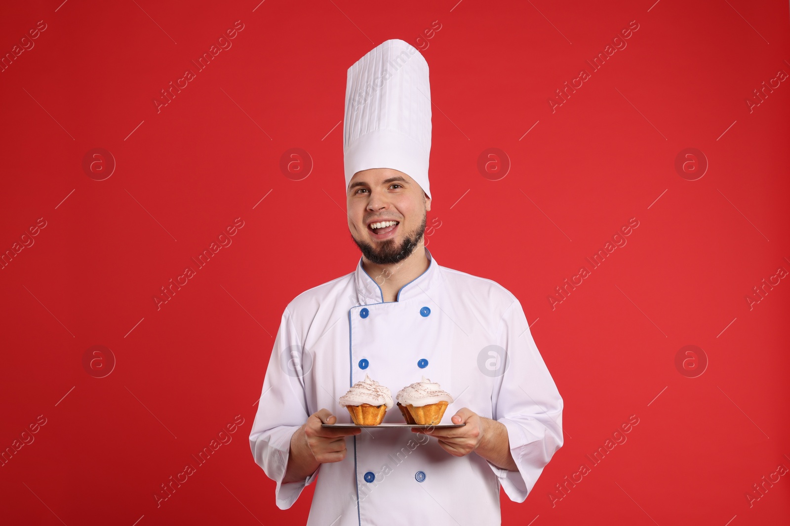 Photo of Happy professional confectioner in uniform holding delicious cupcakes on red background