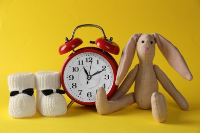 Photo of Alarm clock, bunny and baby booties on yellow background. Time to give birth