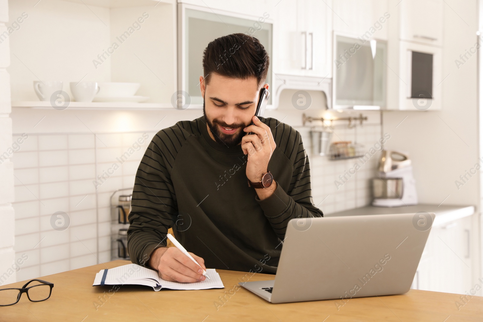 Photo of Handsome young man talking on phone while working at table in kitchen