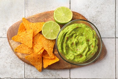 Photo of Wooden board with bowl of delicious guacamole, nachos chips and lime on white tiled table, top view