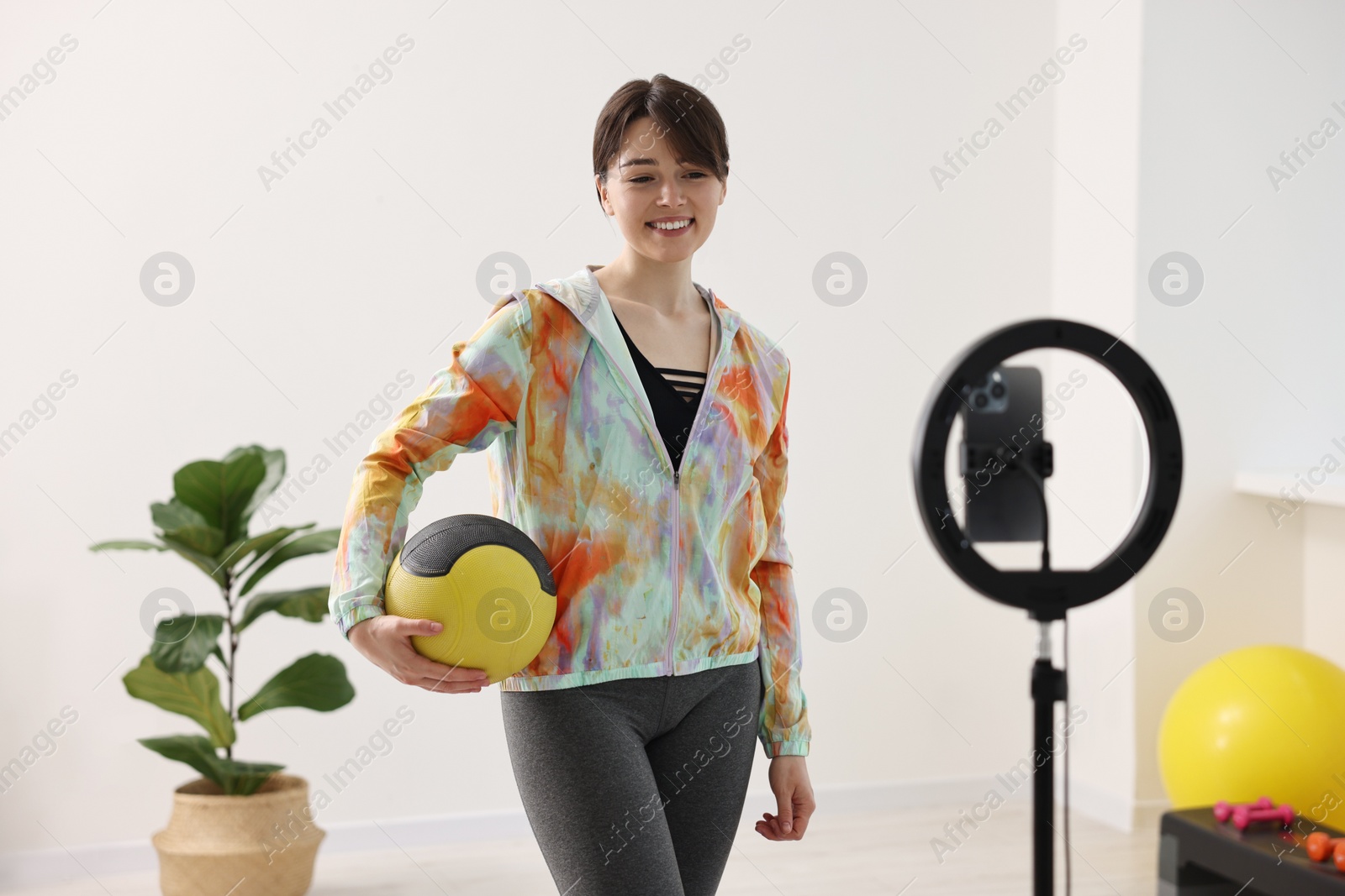 Photo of Happy sports blogger holding medicine ball while streaming online fitness lesson with smartphone at home