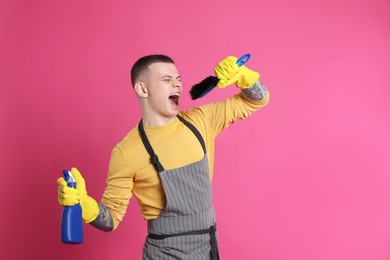 Photo of Handsome young man with brush and bottle of detergent singing on pink background. Space for text