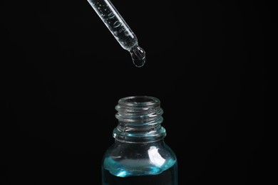 Dripping cosmetic serum from pipette into bottle on black background, closeup