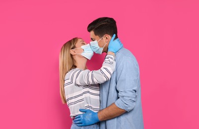 Couple in medical masks and gloves trying to kiss on pink background