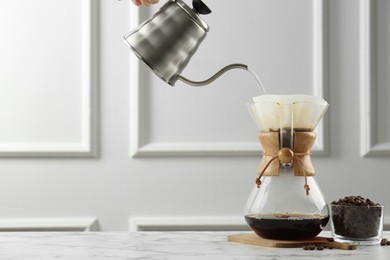 Photo of Pouring hot water from kettle into glass chemex coffeemaker on white marble table, space for text