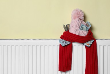 Photo of Knitted hat and scarf with money on heating radiator near beige wall, space for text. Energy crisis concept