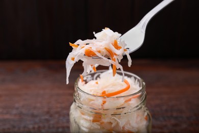 Fork with tasty sauerkraut above glass jar on wooden table, closeup