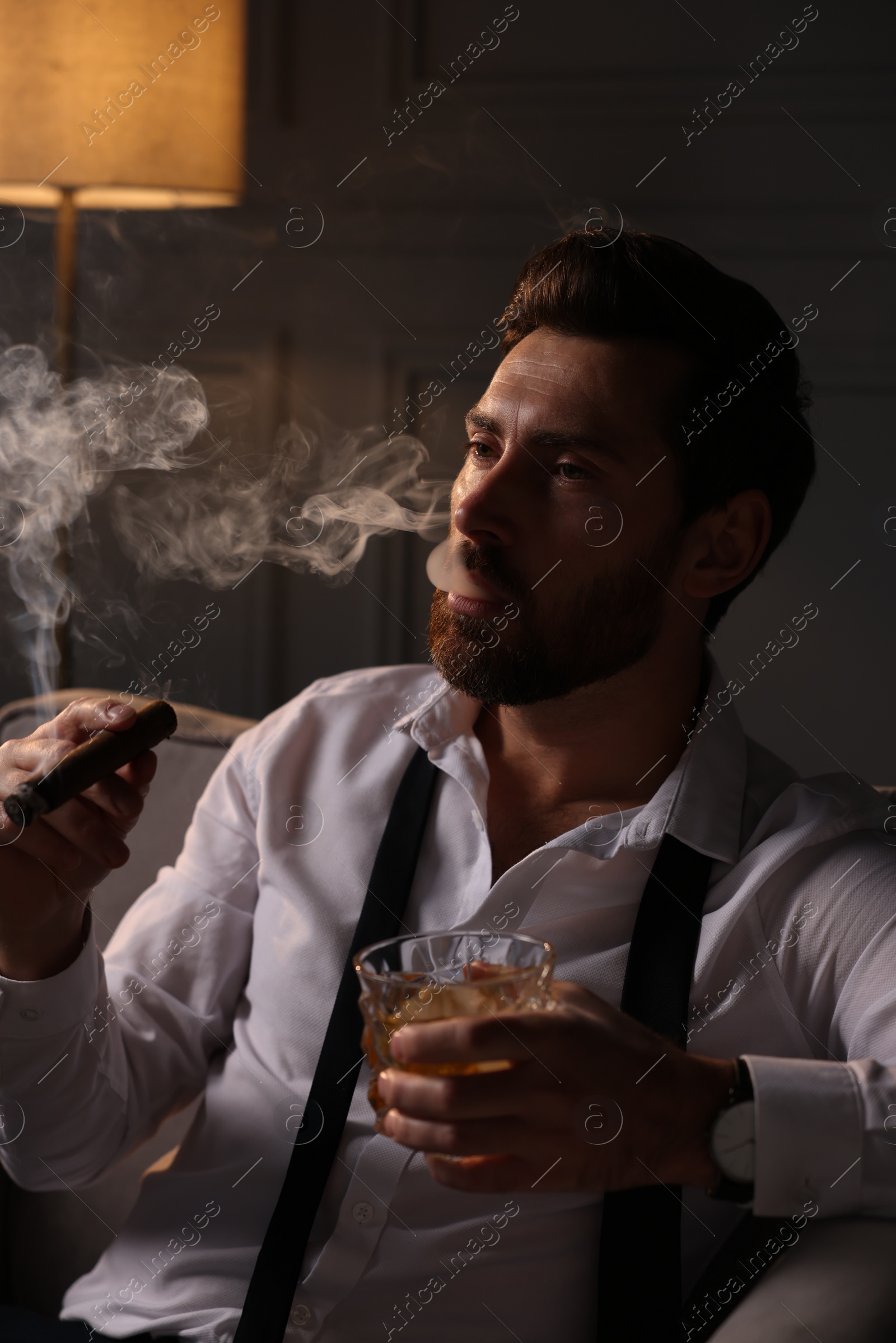 Photo of Handsome man with glass of whiskey smoking cigar in armchair at home