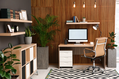 Photo of Comfortable workplace with computer near wooden wall in stylish room interior. Home office design