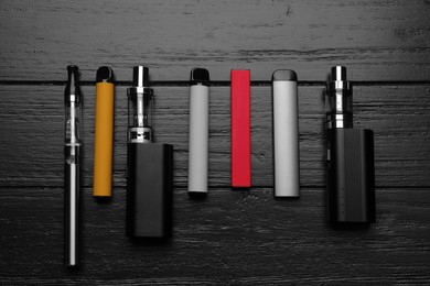 Photo of Group of different electronic cigarettes on black wooden table, flat lay. Smoking alternative
